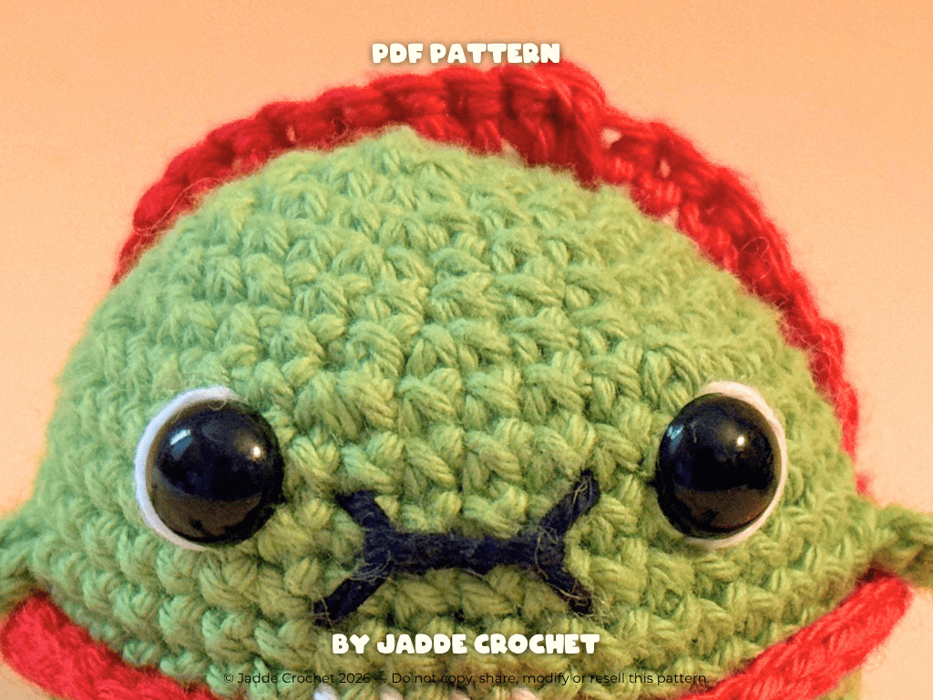Crocheted green character with red hat and black eyes on a peach background, featuring 'Jadde Crochet' branding.