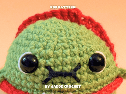 Crocheted green character with red hat and black eyes on a peach background, featuring 'Jadde Crochet' branding.