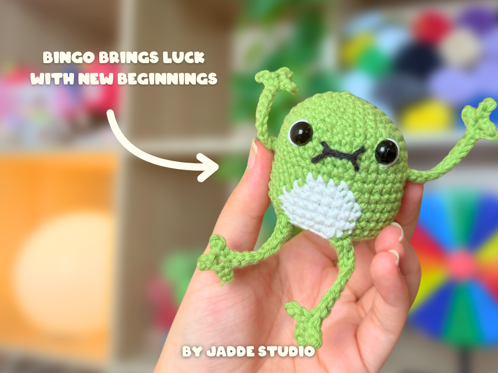 A hand holds a small crocheted frog amigurumi toy with a green body, white belly, and black embroidered eyes and mouth. White text on the image reads 'BINGO BRINGS LUCK WITH NEW BEGINNINGS' and 'BY JADDE STUDIO'.