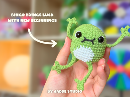 A hand holds a small crocheted frog amigurumi toy with a green body, white belly, and black embroidered eyes and mouth. White text on the image reads 'BINGO BRINGS LUCK WITH NEW BEGINNINGS' and 'BY JADDE STUDIO'.