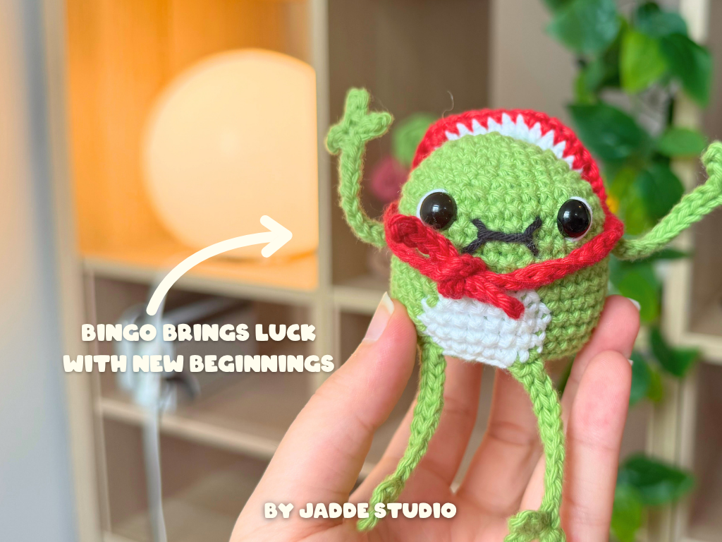 A hand holds a small crocheted frog character wearing a red and white headband and red bow tie. The background is a softly blurred indoor setting with shelves and greenery.