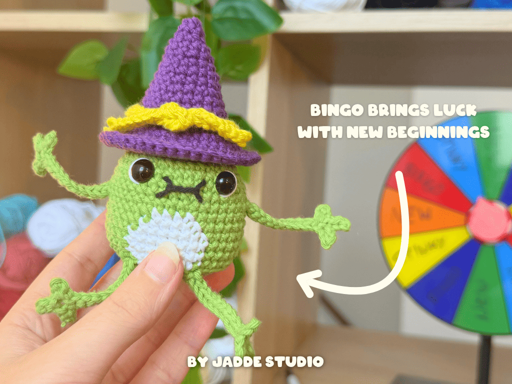 A hand holds a small crocheted black bird character wearing a purple witch hat with a yellow band. The background shows a blurred indoor shelf setting with greenery and colorful items, plus text reading 'KEY BRINGS LUCK FOR PUZZLE SOLVING' and 'BY JADDE STUDIO'.