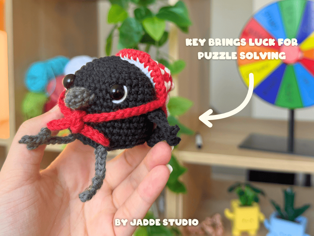 A hand holds a small crocheted black bird character wearing a purple witch hat with a yellow band. The background shows a blurred indoor shelf setting with greenery and colorful items, plus text reading 'KEY BRINGS LUCK FOR PUZZLE SOLVING' and 'BY JADDE STUDIO'.