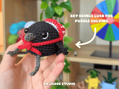 A hand holds a small crocheted black bird character wearing a purple witch hat with a yellow band. The background shows a blurred indoor shelf setting with greenery and colorful items, plus text reading 'KEY BRINGS LUCK FOR PUZZLE SOLVING' and 'BY JADDE STUDIO'.