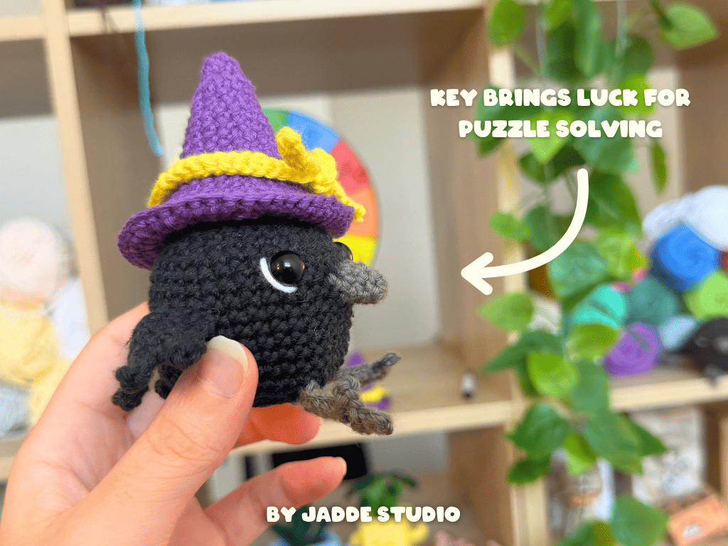 A hand holds a small crocheted black bird character wearing a purple witch hat with a yellow band. The background shows a blurred indoor shelf setting with greenery and colorful items, plus text reading 'KEY BRINGS LUCK FOR PUZZLE SOLVING' and 'BY JADDE STUDIO'.