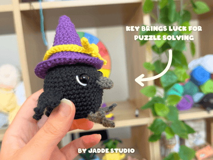 A hand holds a small crocheted black bird character wearing a purple witch hat with a yellow band. The background shows a blurred indoor shelf setting with greenery and colorful items, plus text reading 'KEY BRINGS LUCK FOR PUZZLE SOLVING' and 'BY JADDE STUDIO'.