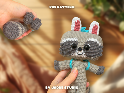A crocheted raccoon doll is shown in a lifestyle setting, held in a hand against a warm, blurred background. Another hand holds a separate crochet arm piece.