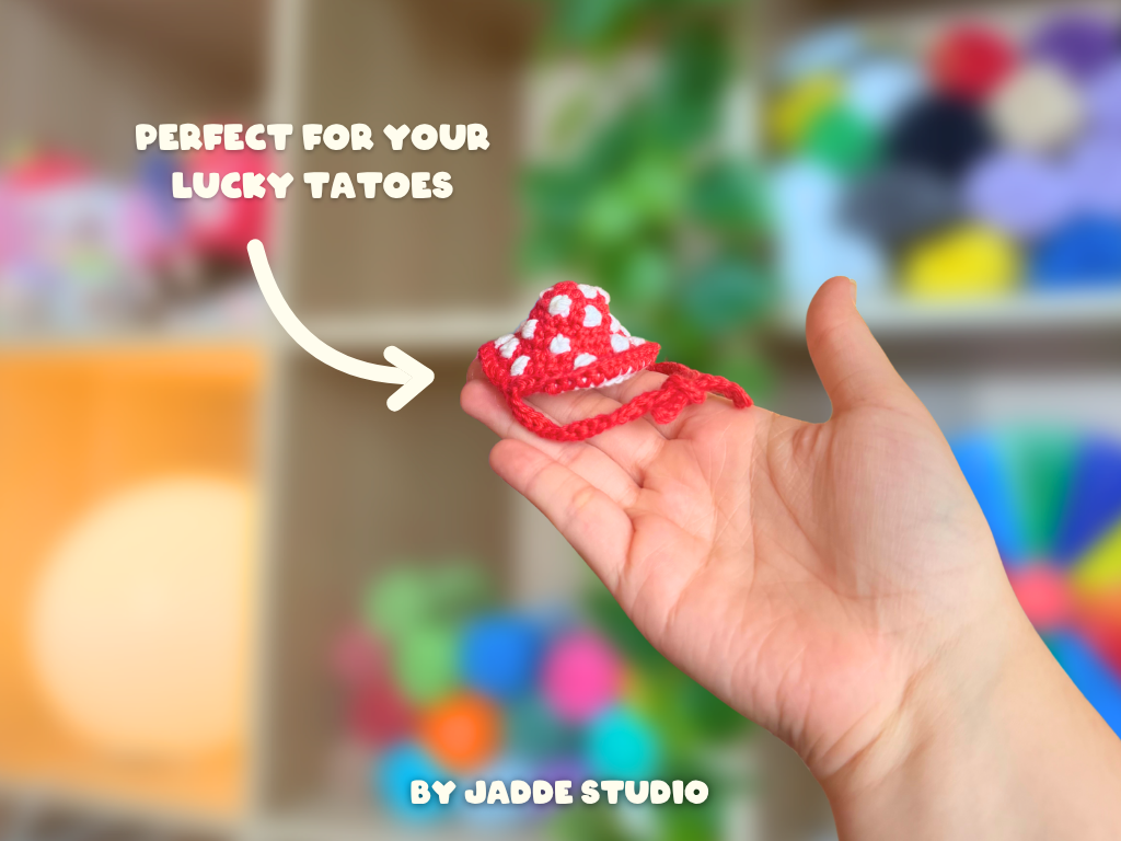 A hand holding a small red and white crocheted lucky charm with white text overlays reading 'PERFECT FOR YOUR LUCKY TATTOES' and 'BY JADDE STUDIO' against a blurred colorful background.