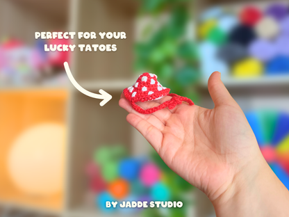 A hand holding a small red and white crocheted lucky charm with white text overlays reading 'PERFECT FOR YOUR LUCKY TATTOES' and 'BY JADDE STUDIO' against a blurred colorful background.