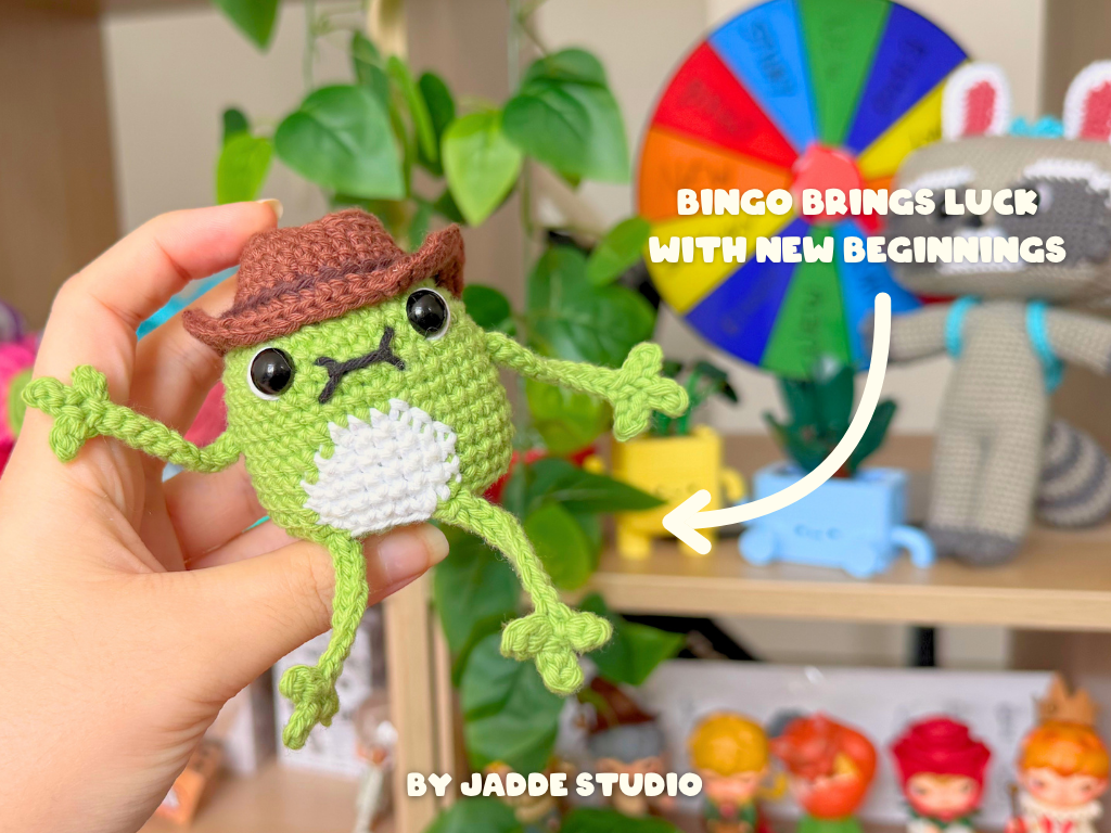 A crocheted frog toy held in a human hand, wearing a brown hat, with a colorful wheel and text overlay in the background. The text reads 'PALOMITA BRINGS GOOD LUCK WITH NEW BEGINNINGS' and 'BY JADDE STUDIO'.