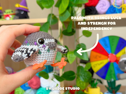 A crocheted owl toy held in a human hand, with orange feet, and a colorful wheel and text overlay in the background. The text reads 'PALOMITA BRINGS LUCK AND STRENGTH FOR INDEPENDENCY' and 'BY JADDE STUDIO'.