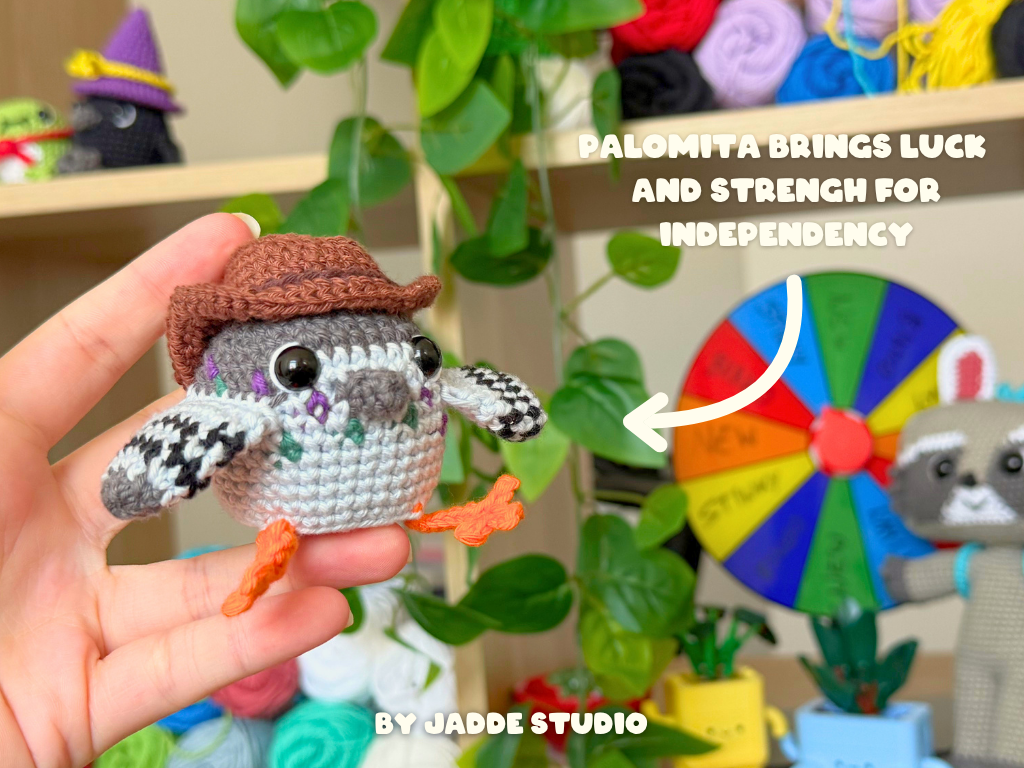 A crocheted pigeon toy held in a human hand, wearing a brown hat and orange feet, with a colorful wheel and text overlay in the background. The text reads 'PALOMITA BRINGS LUCK AND STRENGTH FOR INDEPENDENCY' and 'BY JADDE STUDIO'.