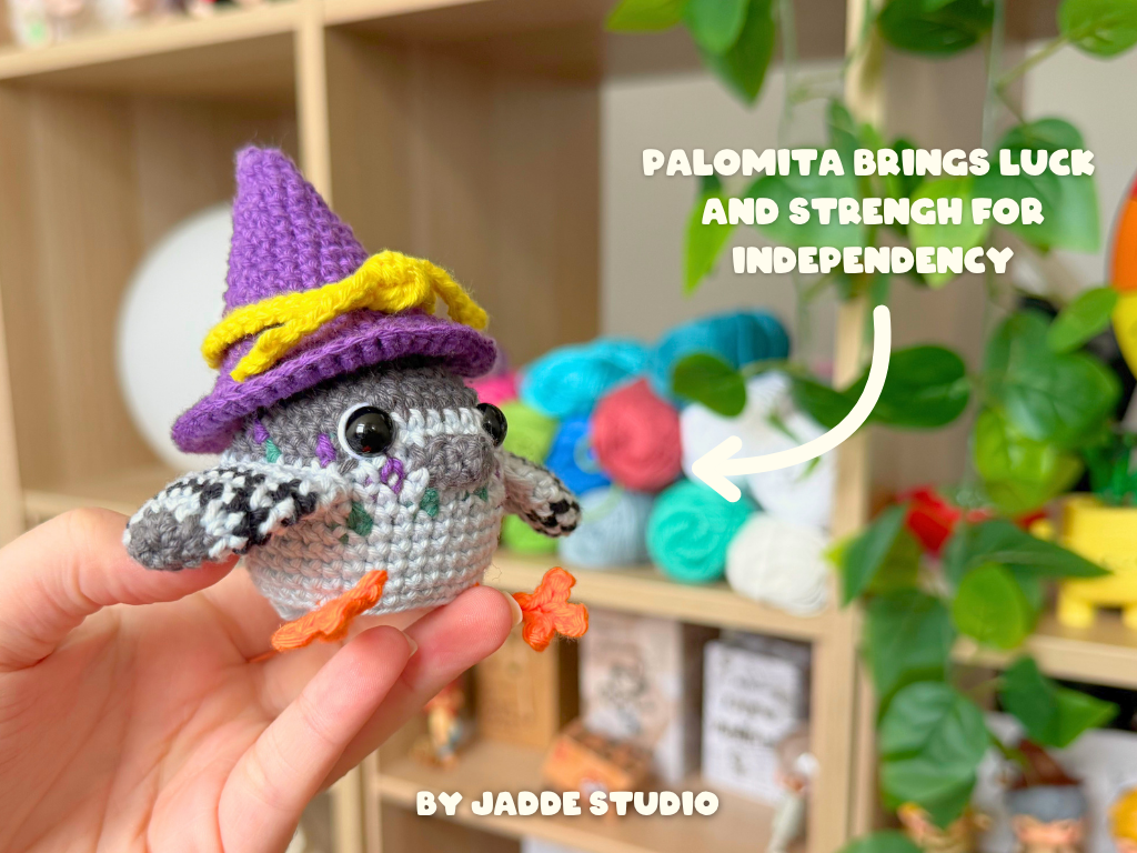 A hand holds a small crocheted bird character wearing a purple witch hat with yellow trim. The background shows a softly blurred indoor shelf setting with colorful yarn balls and green plants.