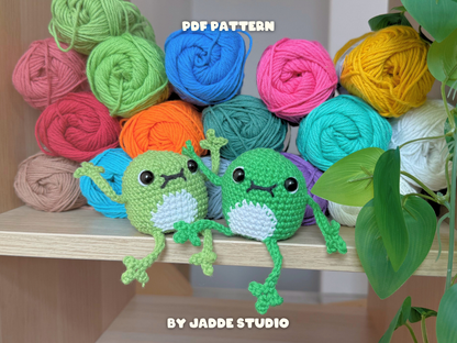 Two green crocheted frog amigurumi toys sitting on a wooden shelf in front of colorful balls of yarn, with text reading 'PDF PATTERN' and 'BY JADDE STUDIO'.