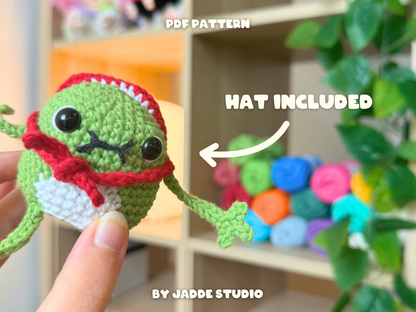 A crocheted green monster amigurumi held in a hand, wearing a red hat and scarf with white belly, with text overlays reading 'PDF PATTERN', 'HAT INCLUDED', and 'BY JADDE STUDIO'.
