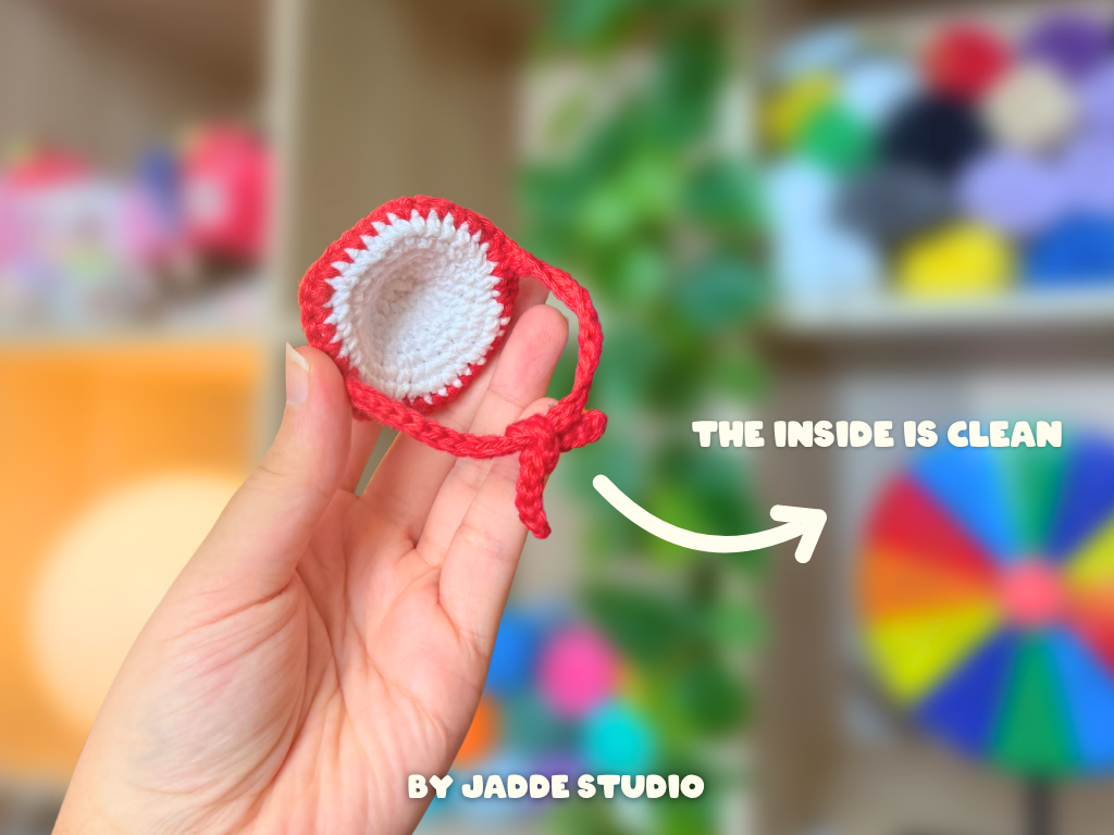A hand holds a small crocheted or knit hat-like accessory in red and white, with text reading 'THE INSIDE IS CLEAN' and 'BY JADDE STUDIO' over a blurred indoor background.