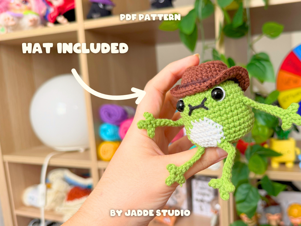 A hand holds a crocheted frog amigurumi in front of a blurred craft room background with shelves of yarn and a rainbow wheel. Text overlays read 'PDF PATTERN', 'HAT INCLUDED' and 'BY JADDE STUDIO'.