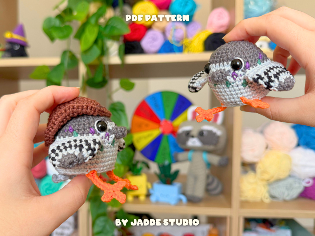 Two hands hold crocheted bird amigurumi toys in front of a blurred craft room background with shelves of yarn and a rainbow wheel. Text overlays read 'PDF PATTERN' and 'BY JADDE STUDIO'.