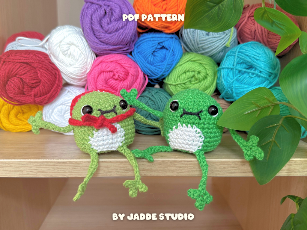 Two crocheted frog amigurumi toys sitting on a wooden shelf in front of colorful balls of yarn, with green leaves in the foreground. Text overlays read 'PDF PATTERN' and 'BY JADDE STUDIO'.