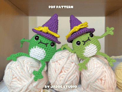 Two crocheted frog characters wearing purple witch hats with yellow bands, posed among large balls of white and pale pink yarn inside a wooden box. Text at the top reads 'PDF PATTERN' and text at the bottom reads 'BY JADDE STUDIO'.
