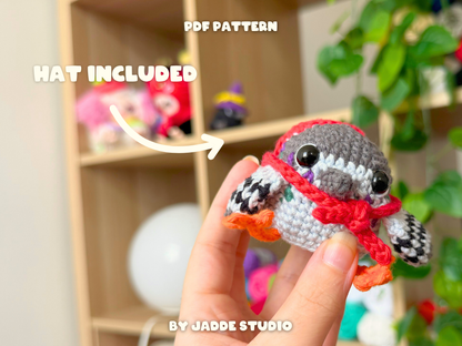 A crocheted bird amigurumi toy is held in a person's hand in front of a blurred shelf display. The bird wears a red hat and has orange feet.