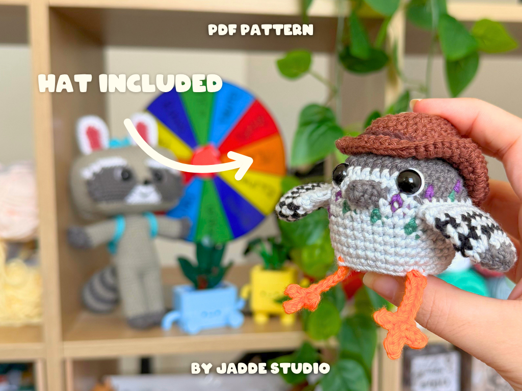 A crocheted bird amigurumi toy is held in a person's hand in front of a blurred shelf display. The bird wears a brown hat and has orange feet.