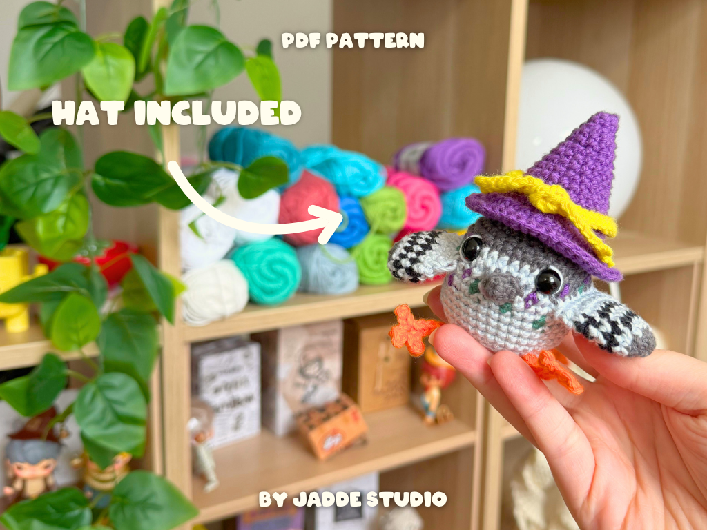 A crocheted bird character toy is held in a person's hand in front of a blurred shelf display. The bird wears a brown hat and has orange feet.