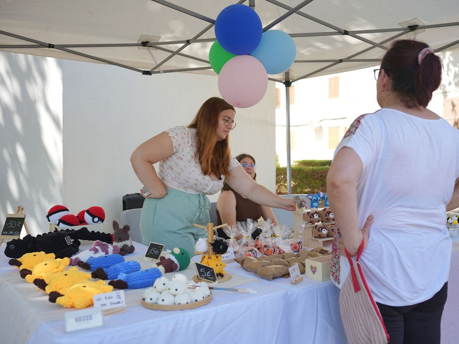 Two women interacting at a table with crochet plushies and various items under a tent with colorful balloons.