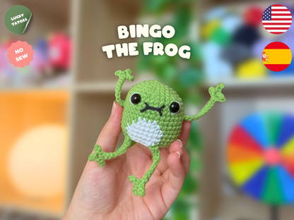 A hand holds a small crocheted frog toy labeled 'BINGO THE FROG' with text overlays including 'LUCKY TATTOOS', 'NO SEW', and country flags. The background is a softly blurred indoor setting with shelves and colorful objects.