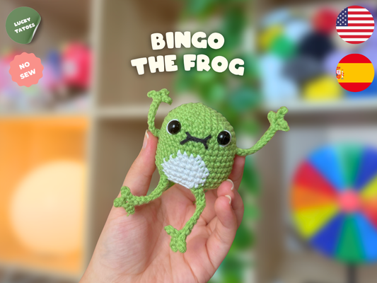 A hand holds a small crocheted frog toy labeled 'BINGO THE FROG' with text overlays including 'LUCKY TATTOOS', 'NO SEW', and country flags. The background is a softly blurred indoor setting with shelves and colorful objects.