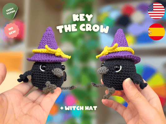 Two small crocheted crow characters wearing purple witch hats with yellow bands are held in human hands against a colorful blurred background. Text reads 'KEY THE CROW' and '+ WITCH HAT', with 'LUCKY TATTOO' and 'NO SEW' badges plus US and Spain flags.