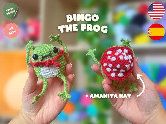 Two hands holding crocheted frog toys in front of a blurred colorful background. The left frog is green with a white belly and red hat, and the right frog has a red with white spots hat.