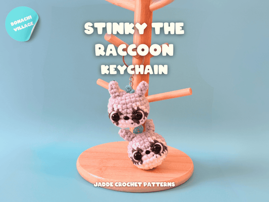 Crocheted raccoon keychain on a stand with 'Stinky the Raccoon' text against a light blue background.