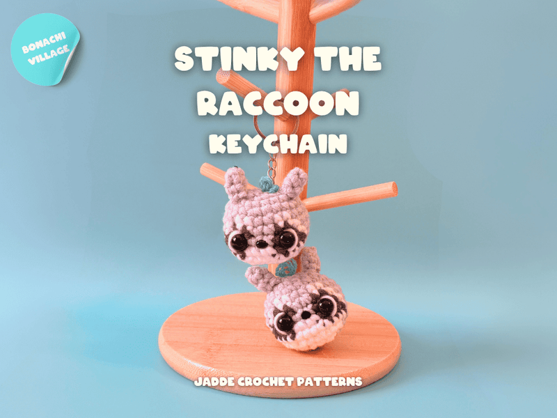 Crocheted raccoon keychain on a stand with 'Stinky the Raccoon' text against a light blue background.