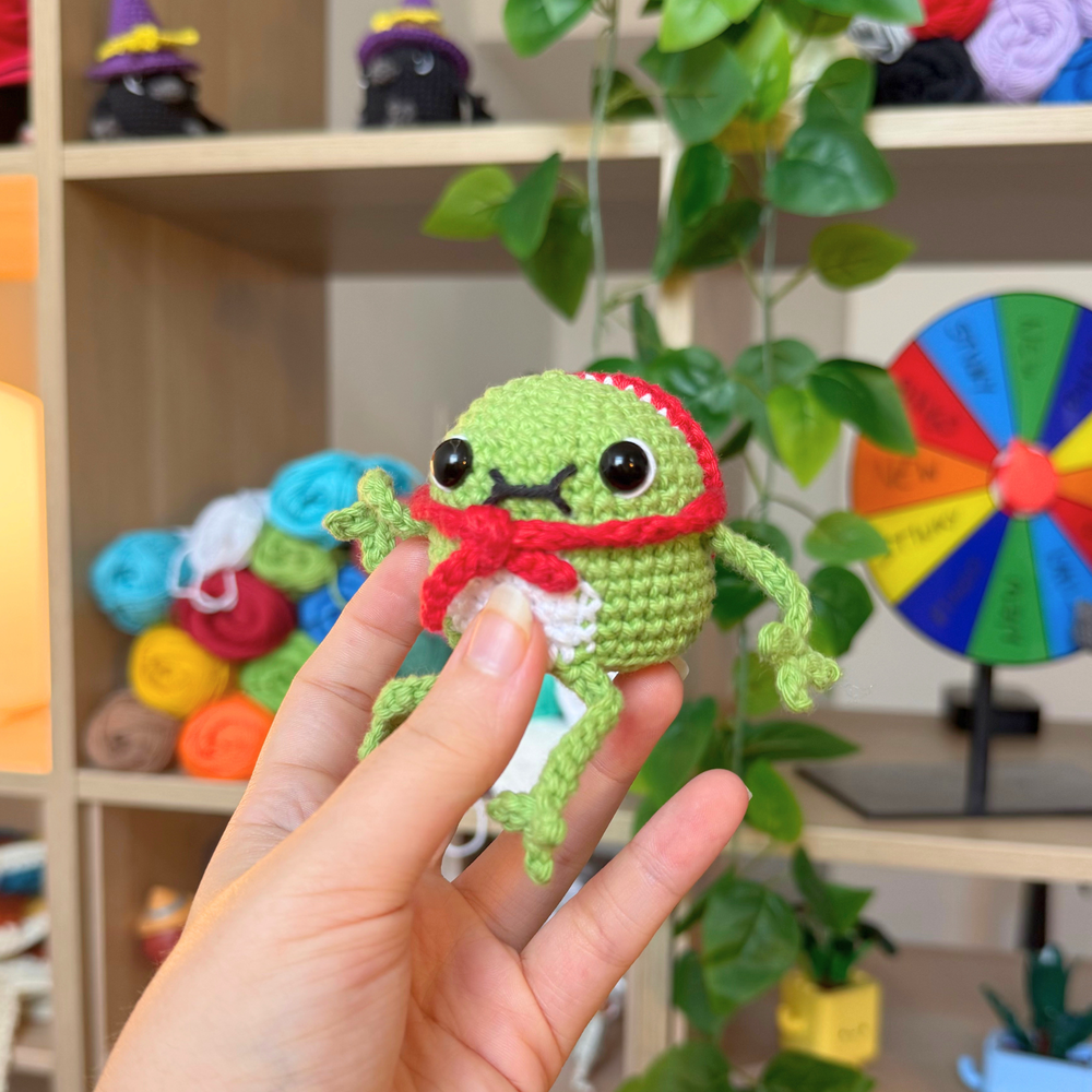 A small crocheted frog toy being held in a person's hand, with a cozy indoor shelf and plant in the background.