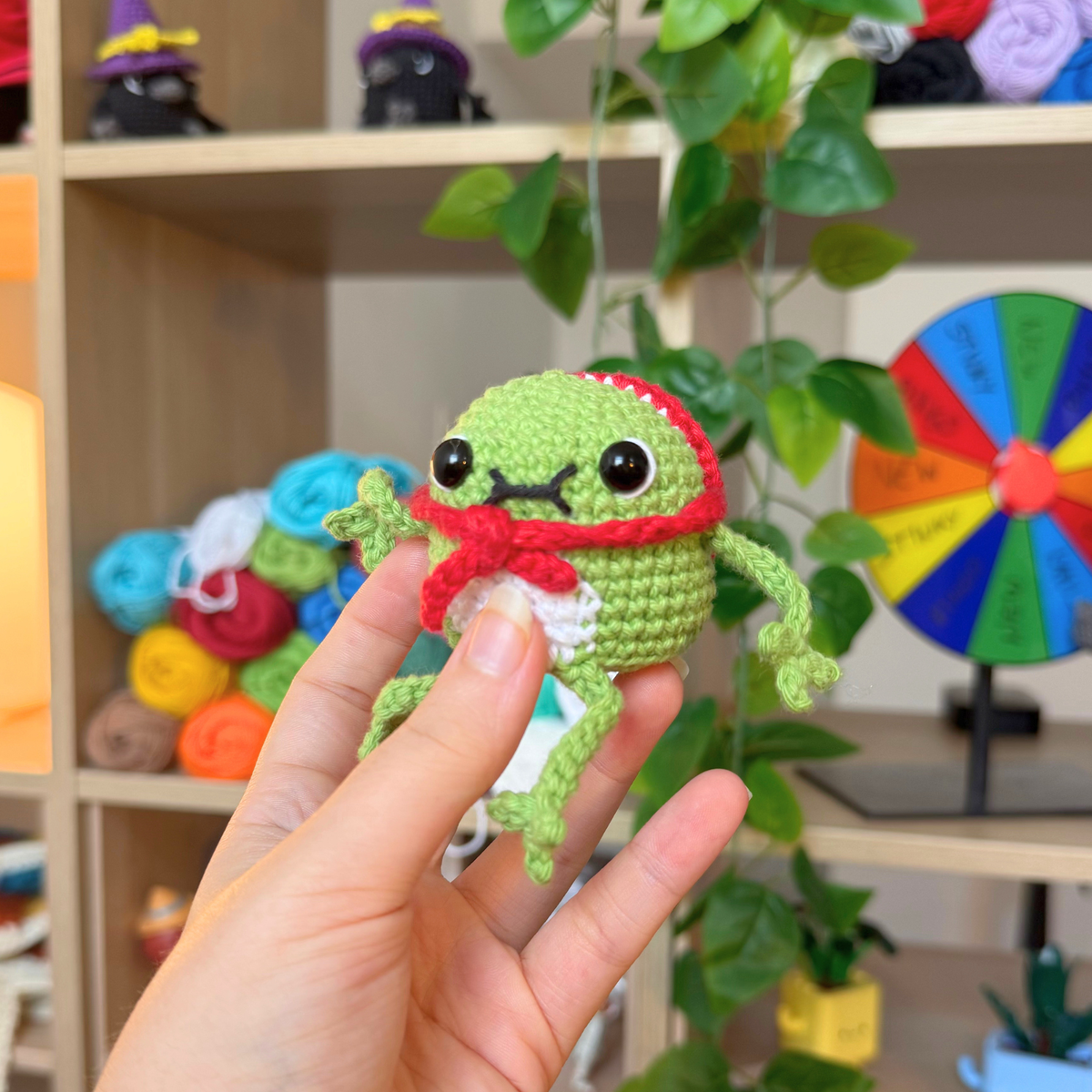 A small crocheted frog toy being held in a person's hand, with a cozy indoor shelf and plant in the background.