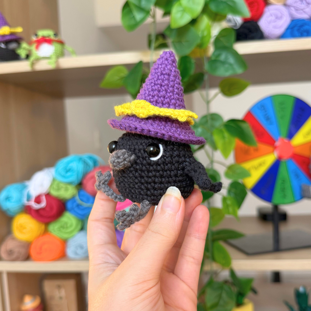 A hand holds a small crocheted black bird character wearing a purple witch hat with a yellow band, set against a cozy craft room background with shelves of yarn and green plants.