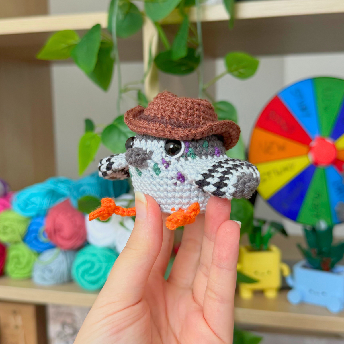 A hand holding a crocheted owl toy wearing a brown hat, with yarn balls, green plants, and a colorful wheel in the background.